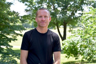 A man, wearing a black t-shirt, poses in front of a small wooded area on the Drillfield. 